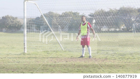 Standing adult male goalkeeper wearing fluorescent bib and red jersey on park pitch, with goal net Standing adult male goalkeeper wearing fluorescent bib and red jersey on park pitch, with goal net 140877919