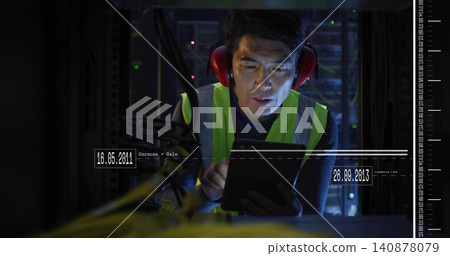 Inspecting tech in hi-vis vest and red muffs, crouching in rack aisle, checking cables using tablet Inspecting tech in hi-vis vest and red muffs, crouching in rack aisle, checking cables using tablet 140878079