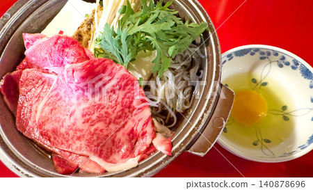 一口盛著大理石紋牛肉和生雞蛋的壽喜燒鍋放在紅色的桌子上。 一口盛著大理石紋牛肉和生雞蛋的壽喜燒鍋放在紅色的桌子上。 140878696