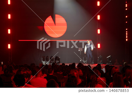 Business speaker presenting on stage to audience at red lit conference with logo projection Business speaker presenting on stage to audience at red lit conference with logo projection 140878853