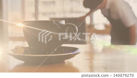 Sitting coffee cup on saucer with biscuit catching flare on wood table in cafe, barista apron Sitting coffee cup on saucer with biscuit catching flare on wood table in cafe, barista apron 140879270