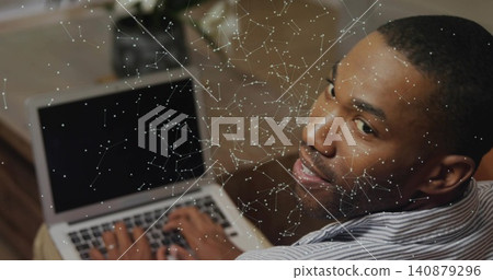 Typing man in light striped shirt turning toward camera at home desk, laptop, plant, copy space Typing man in light striped shirt turning toward camera at home desk, laptop, plant, copy space 140879296
