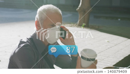 Talking mature man on smartphone holding to-go cup, wearing dark shirt white N95 mask sunlit plaza Talking mature man on smartphone holding to-go cup, wearing dark shirt white N95 mask sunlit plaza 140879409