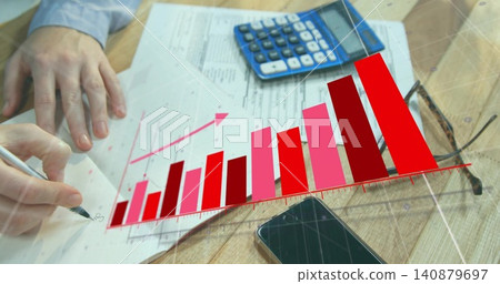 Annotating finance sheets at wooden desk, hands with light blue cuff holding pen and chart overlay 140879697
