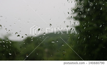 Large raindrops on a windowpane. Through the wet surface, a very blurred silhouette of a green landscape with hills and trees under an overcast sky is visible, creating a melancholy atmosphere. Large raindrops on a windowpane. Through the wet surface, a very blurred silhouette of a green landscape with hills and trees under an overcast sky is visible, creating a melancholy atmosphere. 140880790