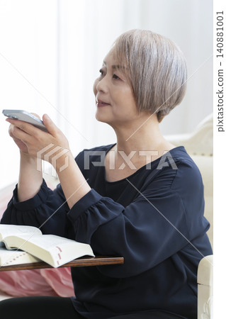 一位老婦人坐在沙發上，用智慧型手機學習英語會話。 140881001