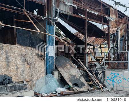 A fire-damaged industrial building exterior featuring collapsed metal structures and charred debris, representing urban decay, catastrophic disaster impact, and architectural ruin A fire-damaged industrial building exterior featuring collapsed metal structures and charred debris, representing urban decay, catastrophic disaster impact, and architectural ruin 140881048