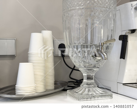 A crystal water dispenser beside white paper cups on a metallic tray in a clean office kitchen, conveying professional hydration services and daily workplace convenience for staff A crystal water dispenser beside white paper cups on a metallic tray in a clean office kitchen, conveying professional hydration services and daily workplace convenience for staff 140881057