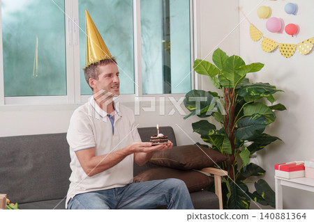 Happy man wearing party hat holding small birthday cake with lit candle on couch Happy man wearing party hat holding small birthday cake with lit candle on couch 140881364