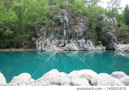 Clear turquoise lake surrounded by lush greenery and rocky cliffs during a sunny day in a tranquil natural setting Clear turquoise lake surrounded by lush greenery and rocky cliffs during a sunny day in a tranquil natural setting 140881678