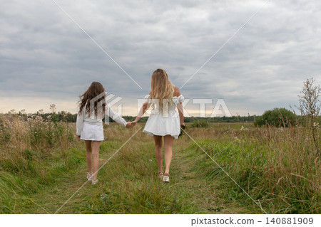 Two girls walking hand-in-hand on a grassy path under a cloudy sky during late afternoon 140881909
