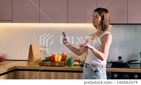 Woman using smartphone to manage grocery order in kitchen. Woman using smartphone to manage grocery order in kitchen. 140882074