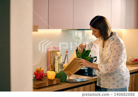 Woman putting fresh lettuce into grocery delivery bag in kitchen. Woman putting fresh lettuce into grocery delivery bag in kitchen. 140882088