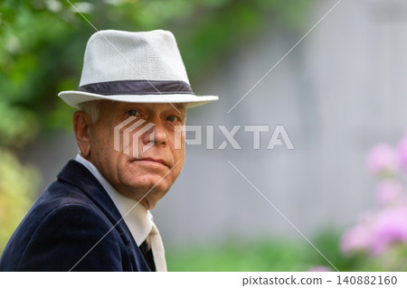 Senior man wearing a white hat and suit sitting outdoors surrounded by greenery and flowers in the summer Senior man wearing a white hat and suit sitting outdoors surrounded by greenery and flowers in the summer 140882160