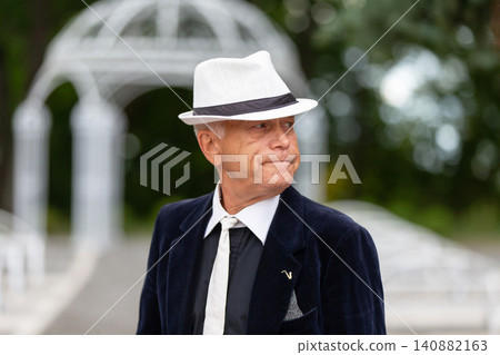 Elderly man in suit and hat poses confidently at outdoor event during daytime, showcasing classic style and elegance Elderly man in suit and hat poses confidently at outdoor event during daytime, showcasing classic style and elegance 140882163