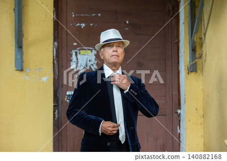An elderly man man adjusts the tie, standing against the yellow wall in the city atmosphere in the daytime An elderly man man adjusts the tie, standing against the yellow wall in the city atmosphere in the daytime 140882168