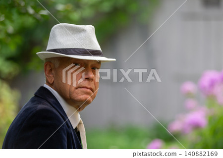 Older man wearing a stylish hat looks thoughtfully at the camera in a vibrant garden setting during a sunny day 140882169
