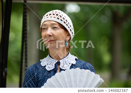Older woman in vintage attire stands with a fan amidst lush greenery in a tranquil outdoor setting 140882387