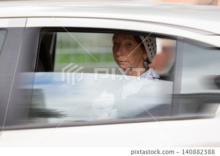 Elderly woman gazes thoughtfully from car window in quiet urban environment during daytime Elderly woman gazes thoughtfully from car window in quiet urban environment during daytime 140882388