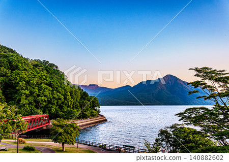 The beautiful surface of Lake Shikotsu, bathed in the soft light of twilight, and the historic red railway bridge of the Yamasen line create a stunning scene. The beautiful surface of Lake Shikotsu, bathed in the soft light of twilight, and the historic red railway bridge of the Yamasen line create a stunning scene. 140882602