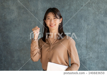 A Japanese woman makes a triumphant gesture while holding a laptop against a gray background. A Japanese woman makes a triumphant gesture while holding a laptop against a gray background. 140883650