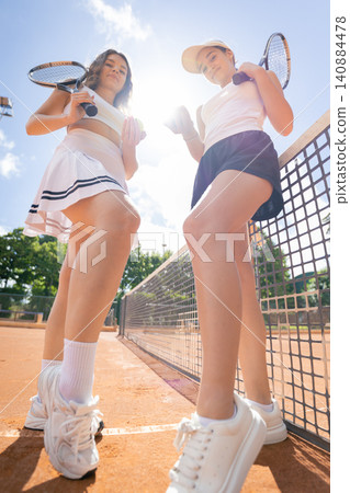 Two young women hold tennis rackets on a court during a sunny day while wearing sports outfits and preparing for a game Two young women hold tennis rackets on a court during a sunny day while wearing sports outfits and preparing for a game 140884478