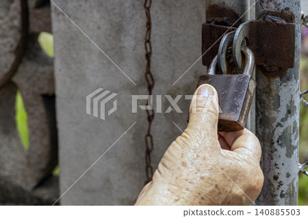 Close-up of Wrinkled Elderly Hand Gripping a Rusty Iron Padlock Attached to Weathered Metal Gate With Hanging Chain, Symbolizing Security, Protection, Aging, and Restricted Access in Rural Environment Close-up of Wrinkled Elderly Hand Gripping a Rusty Iron Padlock Attached to Weathered Metal Gate With Hanging Chain, Symbolizing Security, Protection, Aging, and Restricted Access in Rural Environment 140885503