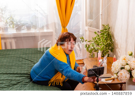 Elderly woman presenting cosmetic favorites while interacting with viewers during a livestream. Beauty blogging and communication concept. Comfortable home setting. Elderly woman presenting cosmetic favorites while interacting with viewers during a livestream. Beauty blogging and communication concept. Comfortable home setting. 140887859