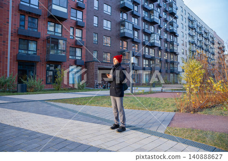 Man standing in modern residential courtyard with smartphone. Hipster guy in red hat using mobile phone near apartment building. Urban lifestyle and digital city communication concept. Man standing in modern residential courtyard with smartphone. Hipster guy in red hat using mobile phone near apartment building. Urban lifestyle and digital city communication concept. 140888627