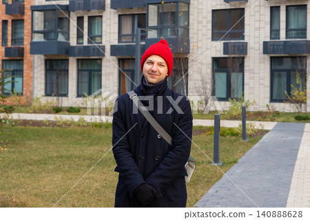Man wearing red beanie and coat standing on street near residential building. Casual outdoor portrait for dating site profile. Male lifestyle and urban city life photography concept. Man wearing red beanie and coat standing on street near residential building. Casual outdoor portrait for dating site profile. Male lifestyle and urban city life photography concept. 140888628