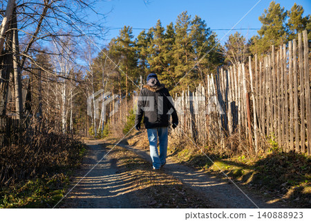 Farmer walking on dirt road in countryside village. Man wearing winter jacket and hat strolling near wooden fence along forest path. Rural lifestyle and agriculture scenery concept. Farmer walking on dirt road in countryside village. Man wearing winter jacket and hat strolling near wooden fence along forest path. Rural lifestyle and agriculture scenery concept. 140888923