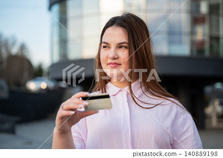 Young woman holding a bank card outside a modern office building during the workday. Online payment and secure digital transactions for everyday cashless shopping Young woman holding a bank card outside a modern office building during the workday. Online payment and secure digital transactions for everyday cashless shopping 140889198