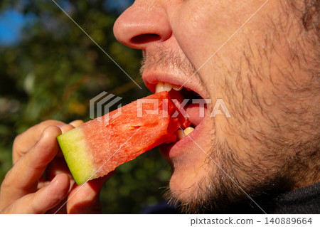 Man eating a fresh slice of juicy watermelon outdoors. Close up of male mouth biting fruit. Healthy summer snack and diet concept. Refreshing red food for a hot weather lifestyle. Man eating a fresh slice of juicy watermelon outdoors. Close up of male mouth biting fruit. Healthy summer snack and diet concept. Refreshing red food for a hot weather lifestyle. 140889664