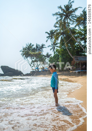5-Year-Old Boy Standing at Shoreline of Sri Lankan Beach, Gentle Waves Lapping 5-Year-Old Boy Standing at Shoreline of Sri Lankan Beach, Gentle Waves Lapping 140889876