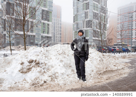Woman standing in snowy city street during winter. Casual girl posing near large snow drift against background of residential high-rise buildings. Urban lifestyle and cold weather season. Woman standing in snowy city street during winter. Casual girl posing near large snow drift against background of residential high-rise buildings. Urban lifestyle and cold weather season. 140889991