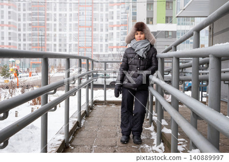 Woman in winter clothes standing on ramp outside building. Girl wearing fur hat and puffer jacket on cold day. Urban residential area view during snowfall. Seasonal lifestyle portrait. Woman in winter clothes standing on ramp outside building. Girl wearing fur hat and puffer jacket on cold day. Urban residential area view during snowfall. Seasonal lifestyle portrait. 140889997