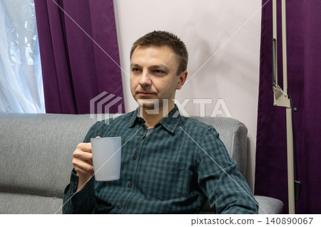 Man sitting on a sofa and holding a cup of hot drink. Relaxed guy resting at home during break. Peaceful leisure time and daily lifestyle of a young person in indoor environment. Man sitting on a sofa and holding a cup of hot drink. Relaxed guy resting at home during break. Peaceful leisure time and daily lifestyle of a young person in indoor environment. 140890067