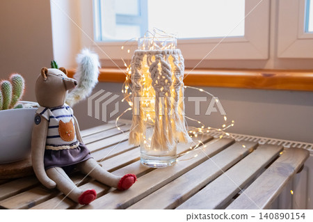 Macrame glass lantern with glowing fairy lights on wooden table. Soft home decor with handmade textile craft and fabric doll. Cozy atmosphere for interior design and relaxation. Macrame glass lantern with glowing fairy lights on wooden table. Soft home decor with handmade textile craft and fabric doll. Cozy atmosphere for interior design and relaxation. 140890154