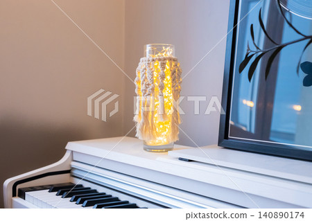 Macrame lamp with fairy light in glass vase. Handmade home decor item sitting on white piano. Interior design accessory for cozy atmosphere and soft warm glow in living room living space. Macrame lamp with fairy light in glass vase. Handmade home decor item sitting on white piano. Interior design accessory for cozy atmosphere and soft warm glow in living room living space. 140890174
