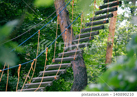 Rope park suspension bridge with dark wooden steps in summer forest Rope park suspension bridge with dark wooden steps in summer forest 140890210