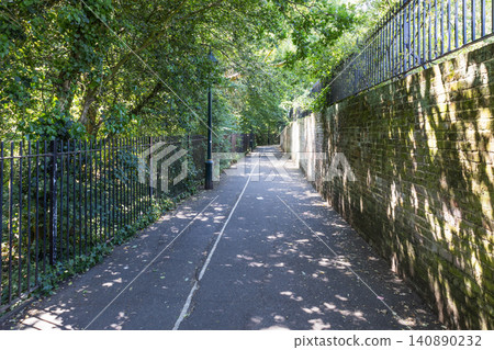 Paved pedestrian path beside a stone wall and lush green trees in a park Paved pedestrian path beside a stone wall and lush green trees in a park 140890232