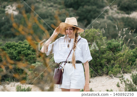 Young woman in white outfit and straw hat enjoying nature walk in scenic landscape 140892250