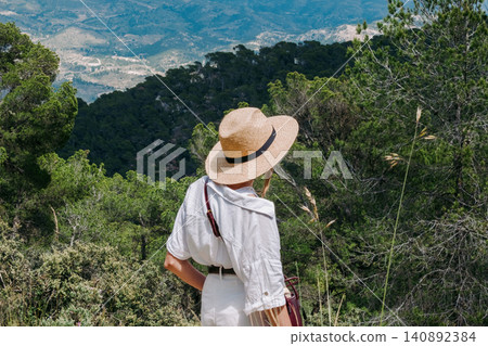 Woman in straw hat enjoying scenic mountain view surrounded by lush green forest Woman in straw hat enjoying scenic mountain view surrounded by lush green forest 140892384