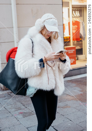 Fashionable young woman in winter wear browsing phone outdoors Fashionable young woman in winter wear browsing phone outdoors 140892876