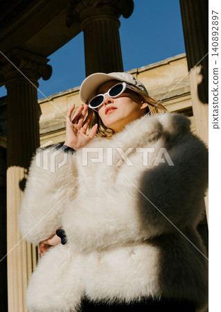 Stylish woman in fur coat and sunglasses posing outside historic building on sunny day 140892897