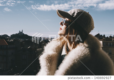 Trendy woman in sunglasses and fur coat enjoying sunny cityscape with blue skies 140892927