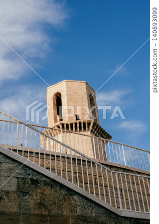 Historic stone tower against blue sky behind modern white fence architecture 140893099