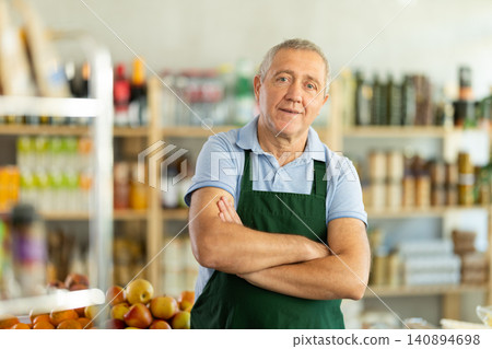 Elderly man in green apron stands near counter, works as salesman in grocery store. Elderly man in green apron stands near counter, works as salesman in grocery store. 140894698