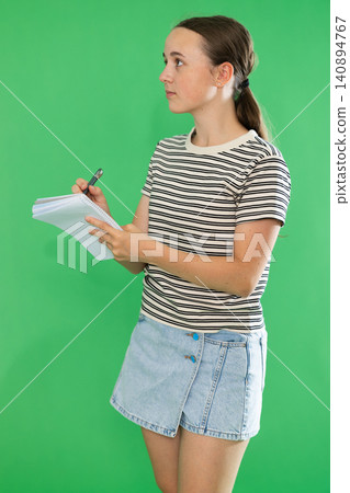 Teen elegant girl dressed in summer T-shirt and blue jeans skirt posing against green backdrop holding documents or writing notes in notebook 140894767
