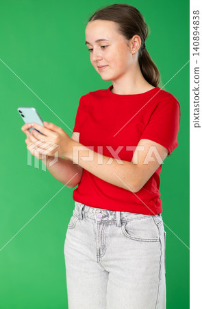 Portrait of positive teen girl dressed in red T-shirt and blue jeans scrolling smartphone standing against green screen Portrait of positive teen girl dressed in red T-shirt and blue jeans scrolling smartphone standing against green screen 140894838